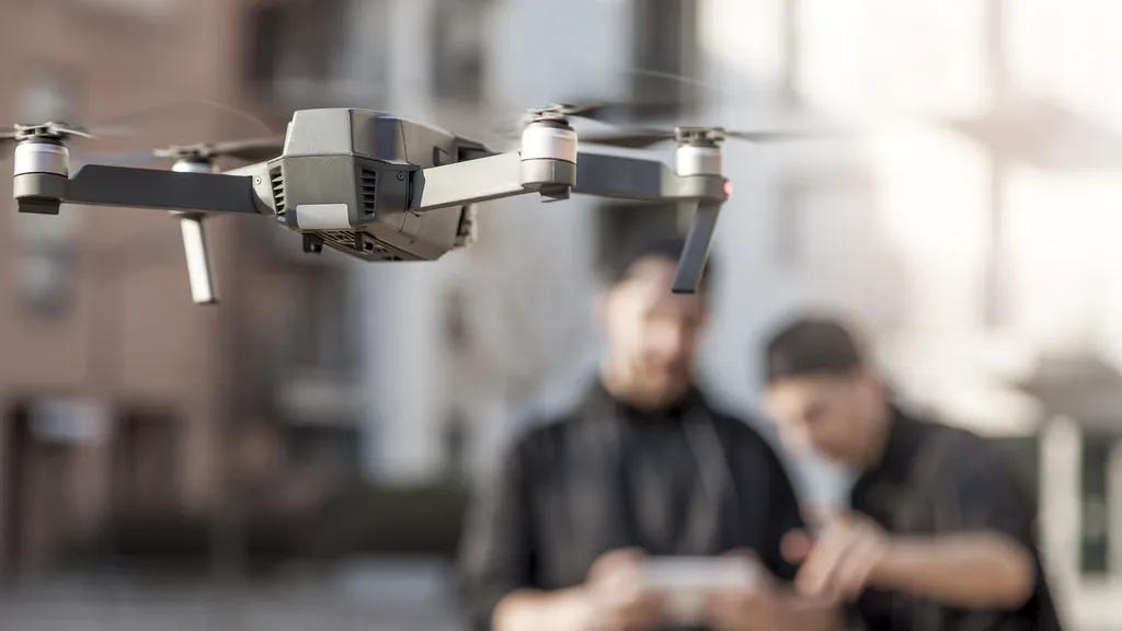 A camera drone hovering in midair while two people operate it from the ground behind it.