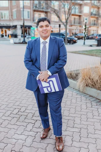 David Mendez in a navy suit, standing on a brick sidewalk in front of a multi-story residential building and holding a Realproz-branded folder.